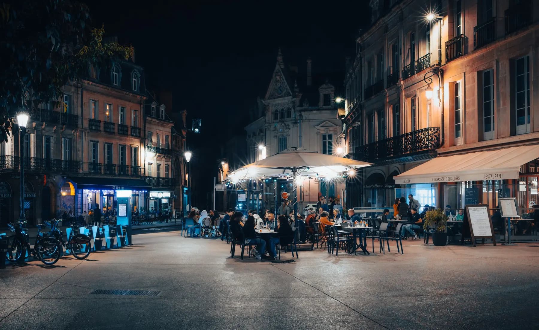 Une place de centre-ville, la nuit : les terrasses d’un bar, d’un café et d’une brasserie sont pleines.